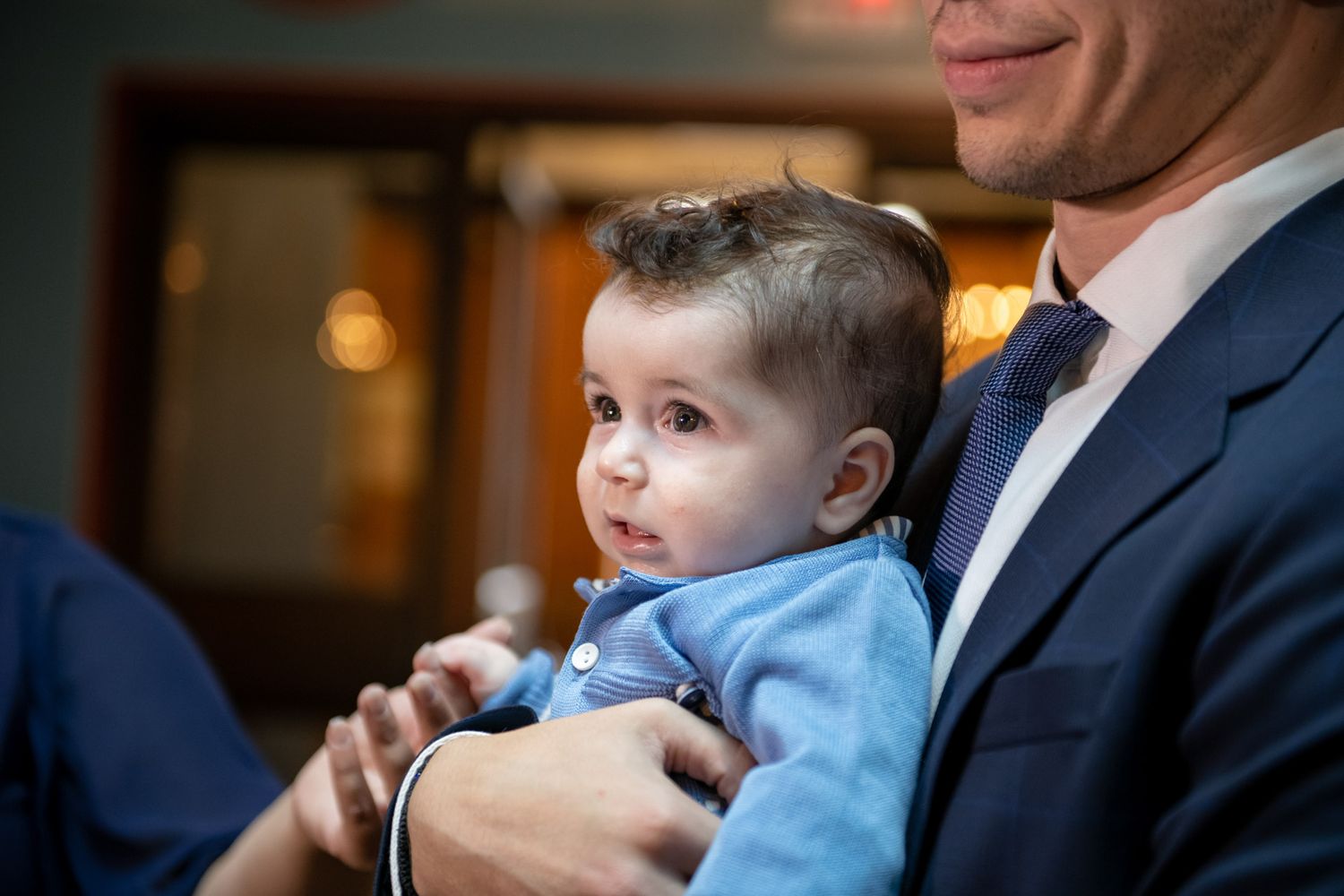 Baby in blue outfit being held by man in suit at Orthodox baptism ceremony.