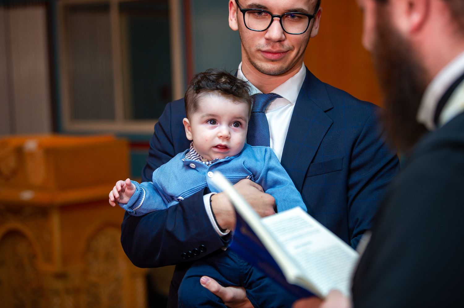 Man in suit holding baby while priest reads from book during Orthodox baptism.
