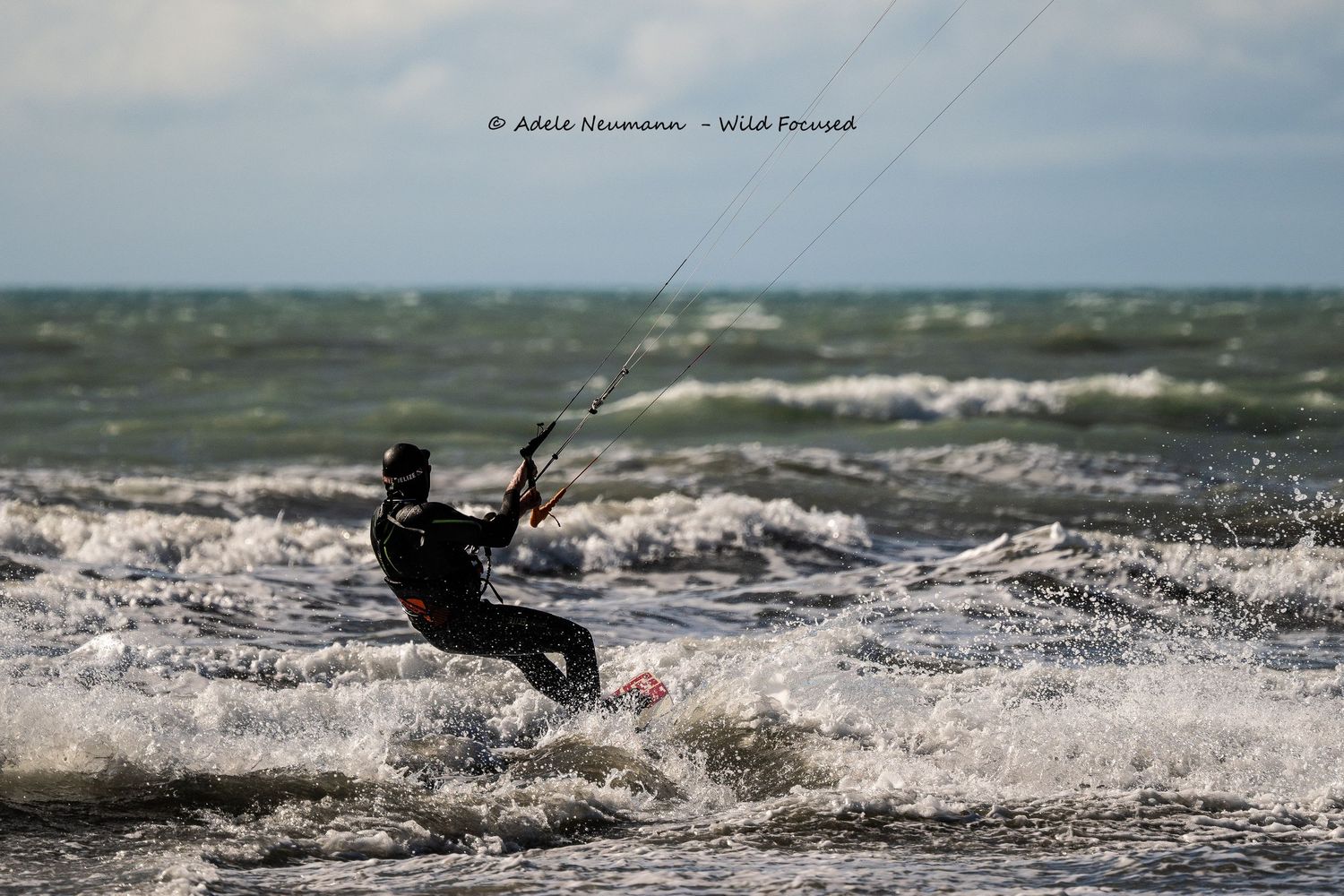 Kitesurfer riding waves in choppy ocean waters wearing a black wetsuit with cloudy sky.