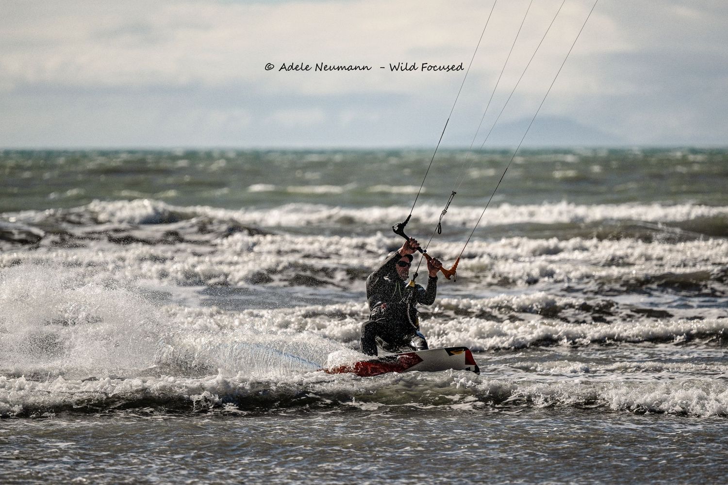 A kitesurfer rides powerful ocean waves in choppy conditions, controlling a large kite against a cloudy sky.
