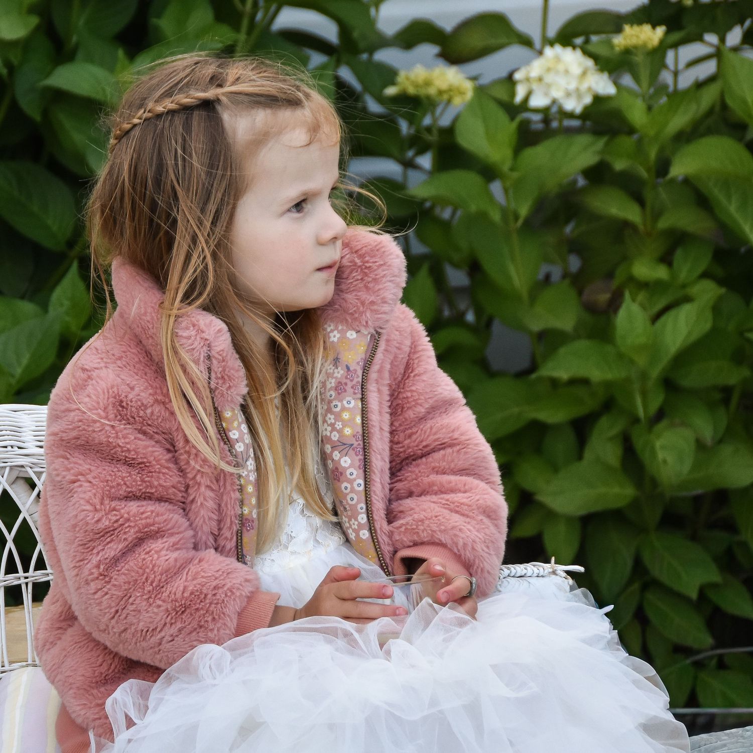 Young girl in pink fluffy jacket and white tulle dress sits outdoors near hydrangea bushes.