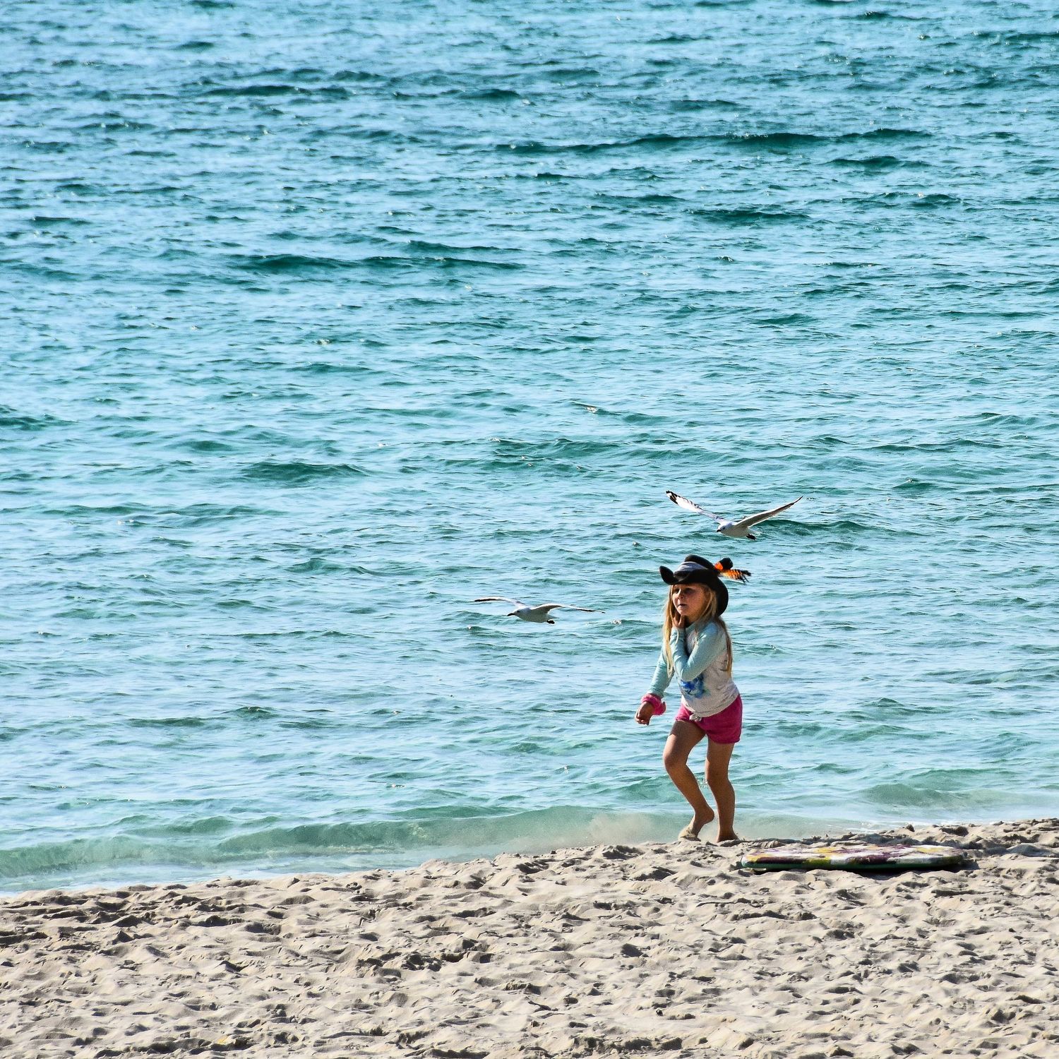 A young girl runs along a sandy beach near turquoise ocean waves, as two seagulls fly overhead on a sunny day.