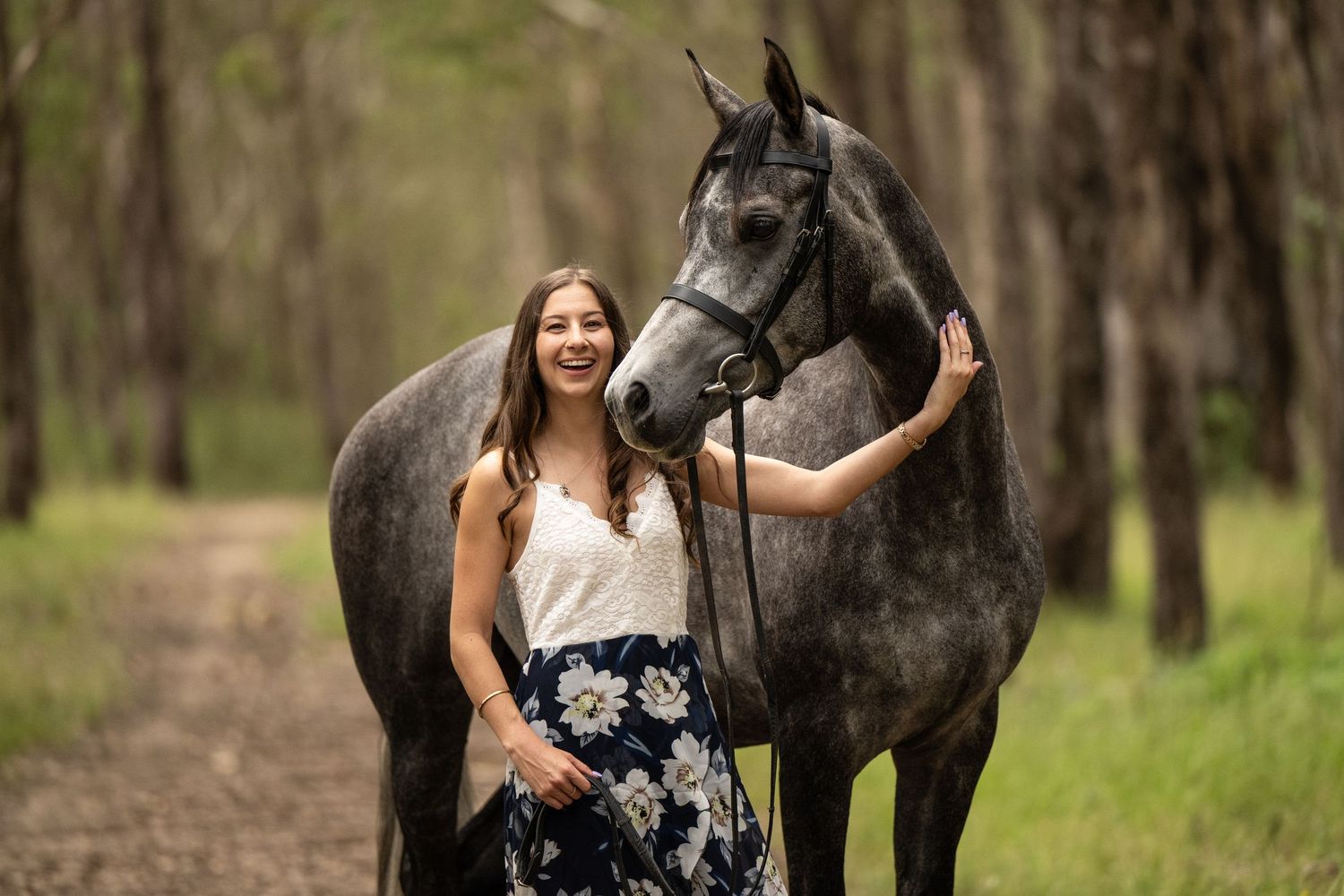 Young woman in floral skirt smiling beside a dapple grey horse on a forest trail.