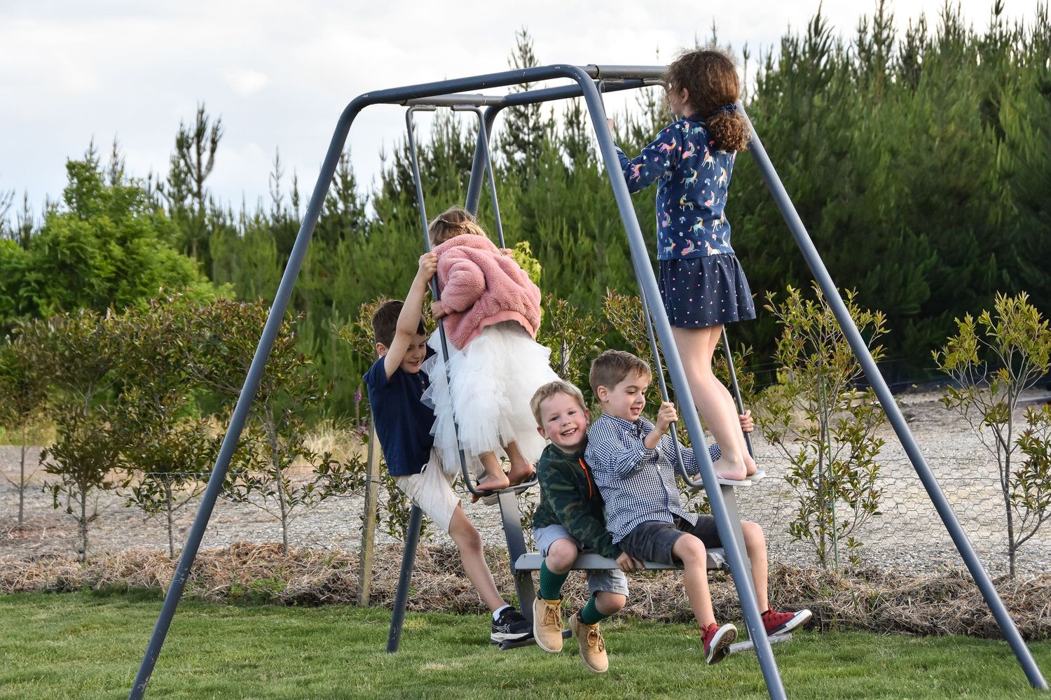 Children playing on a metal swing set in a garden with trees in the background.