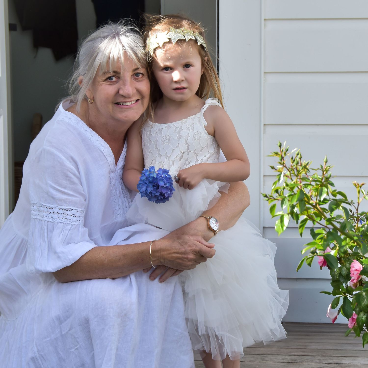 Woman in white dress hugging young girl in white dress with blue hydrangea flower outdoors.