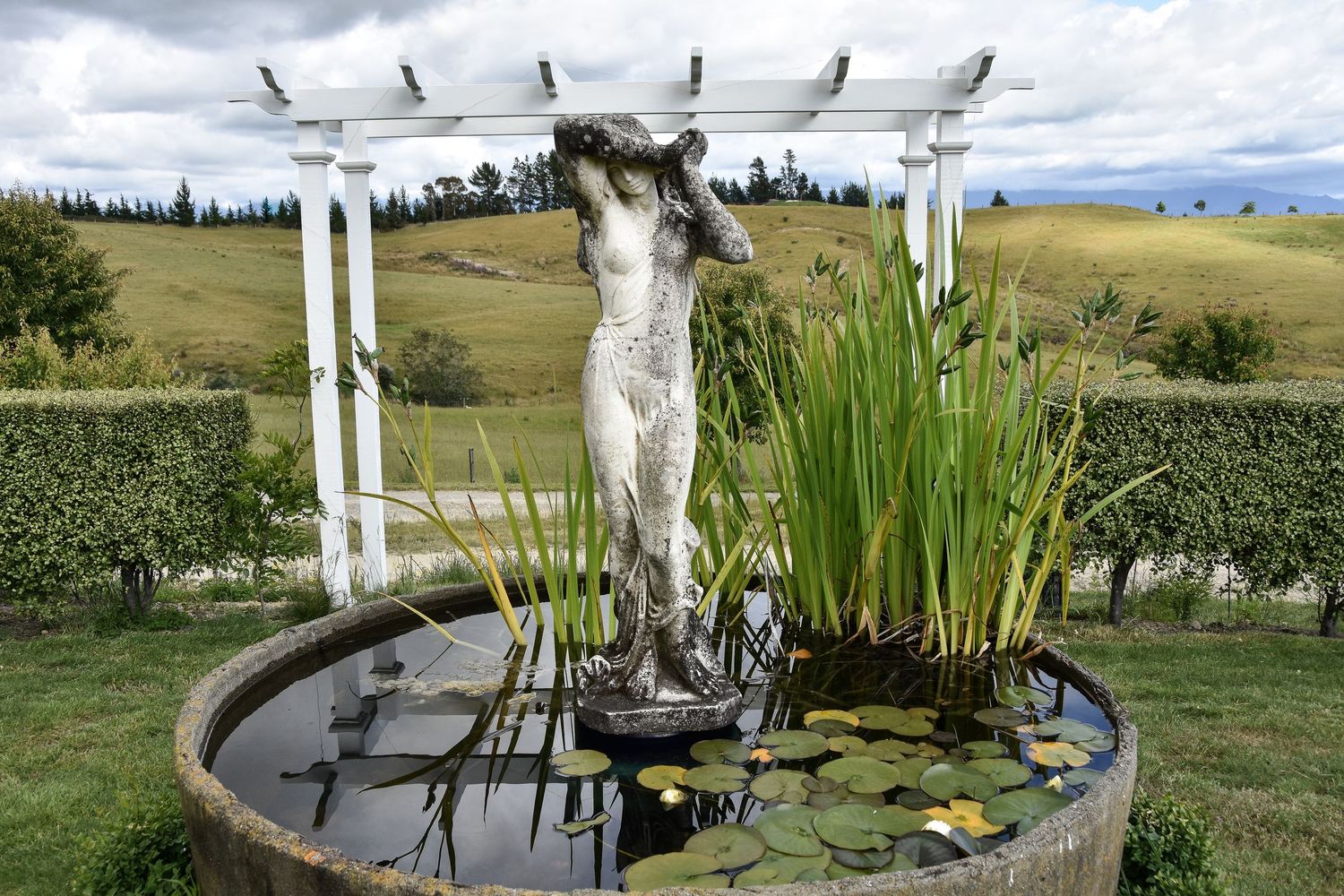 Stone statue of a figure in a round garden pond with lily pads and reeds, white pergola and rolling hills behind.