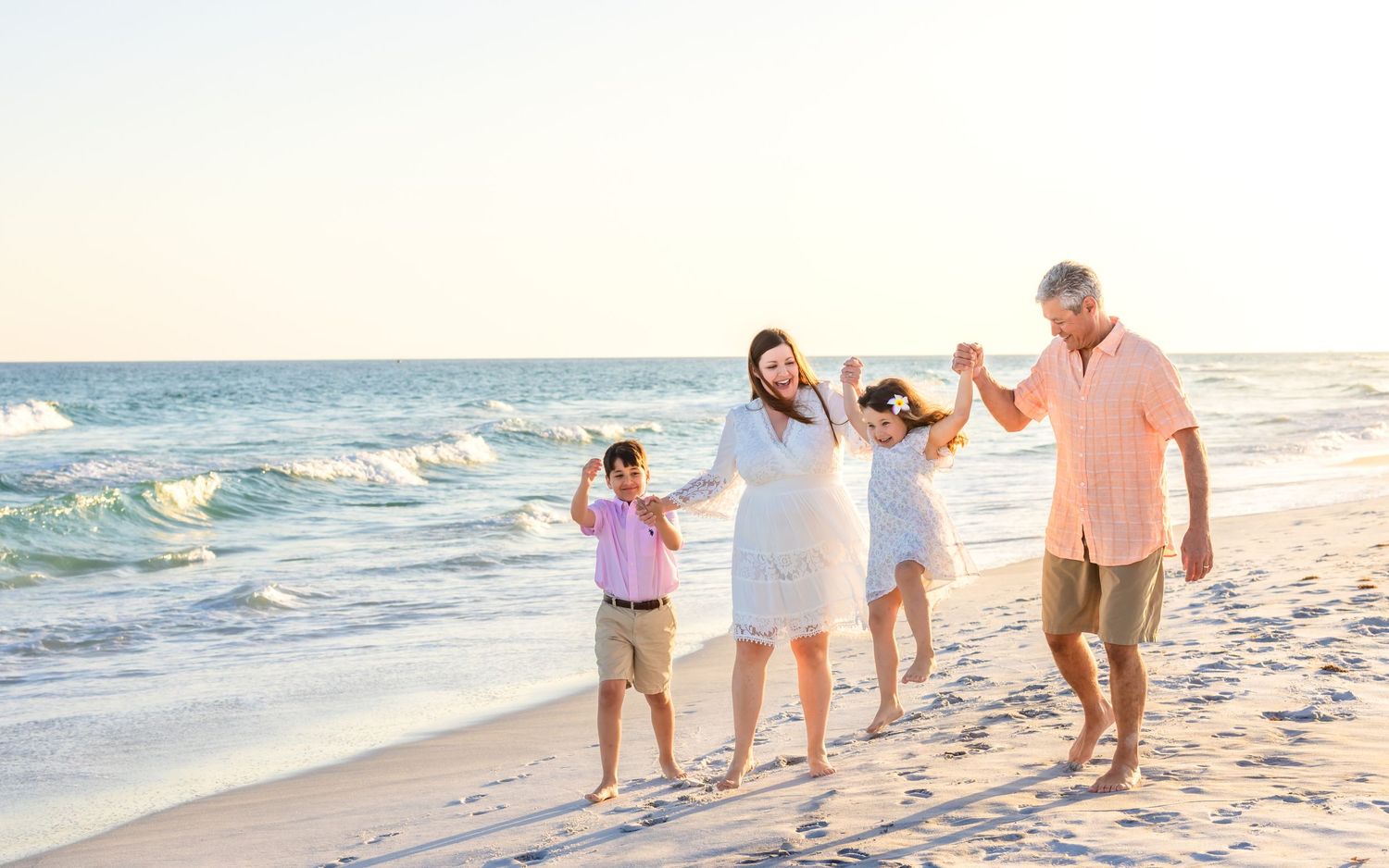 Happy family of four walking hand-in-hand on a sunny beach, with ocean waves in the background.