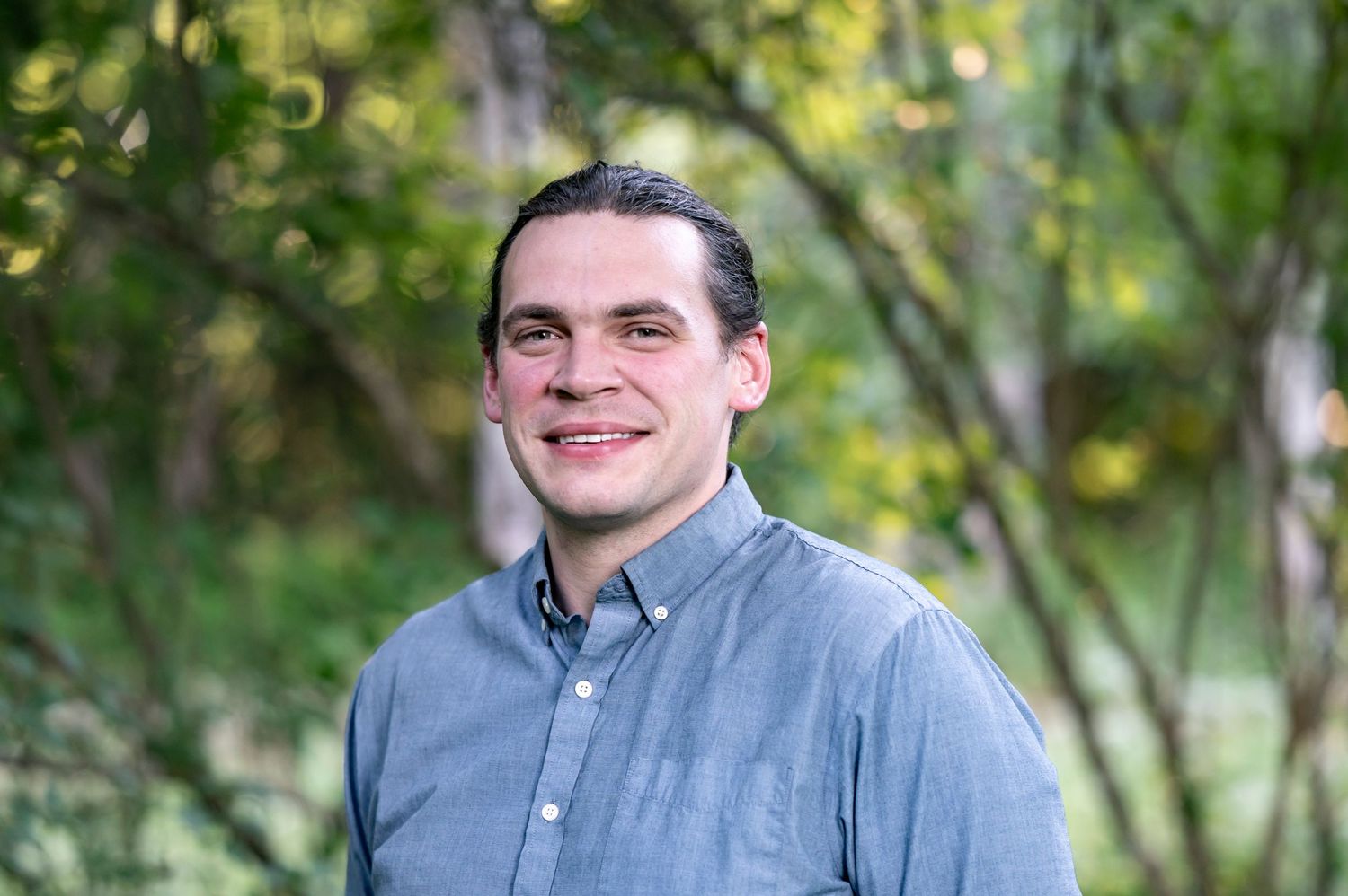 Smiling young man in a blue shirt outdoors with green trees in the background.