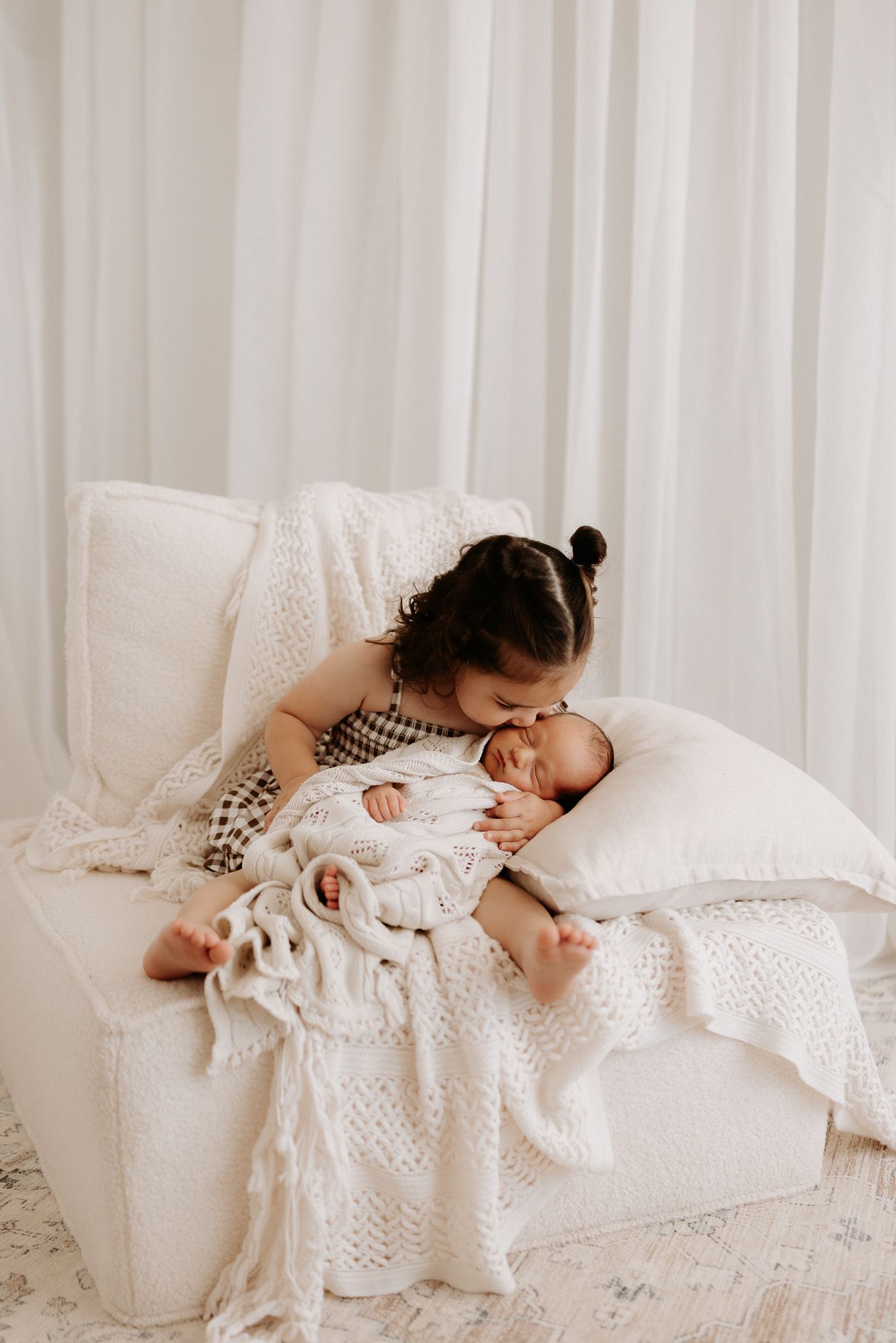 Newborn baby sleeping peacefully on white cushion wrapped in cream knit outfit surrounded by soft white blankets.