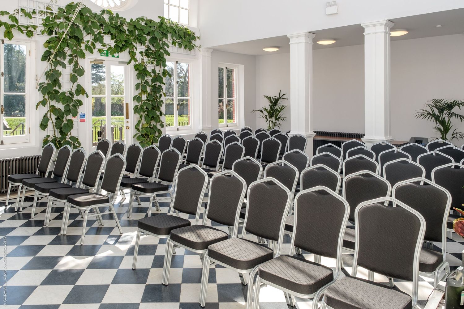 Elegant event room with black and white checkered floors, rows of banquet chairs, white columns, and lush green ivy decor.