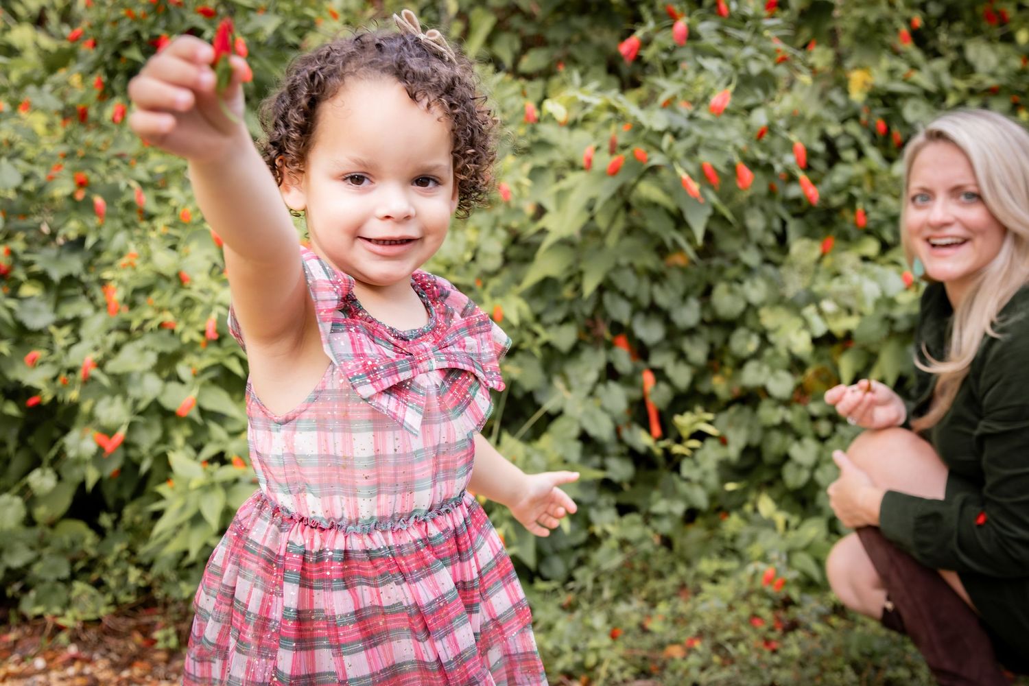 A blonde woman crouching to high-five a curly-haired toddler girl in a plaid dress by a wooden fence outdoors.
