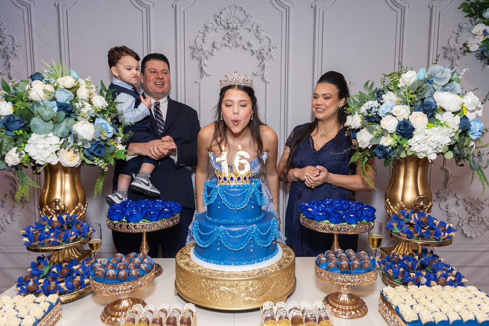 Family celebrating a sweet 16 birthday party with a blue tiered cake, gold accents, and floral arrangements.