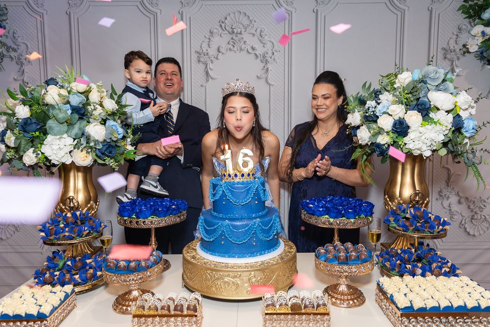 Family celebrating a girl's 16th birthday with a blue tiered cake, cupcakes, and floral arrangements at an elegant party.