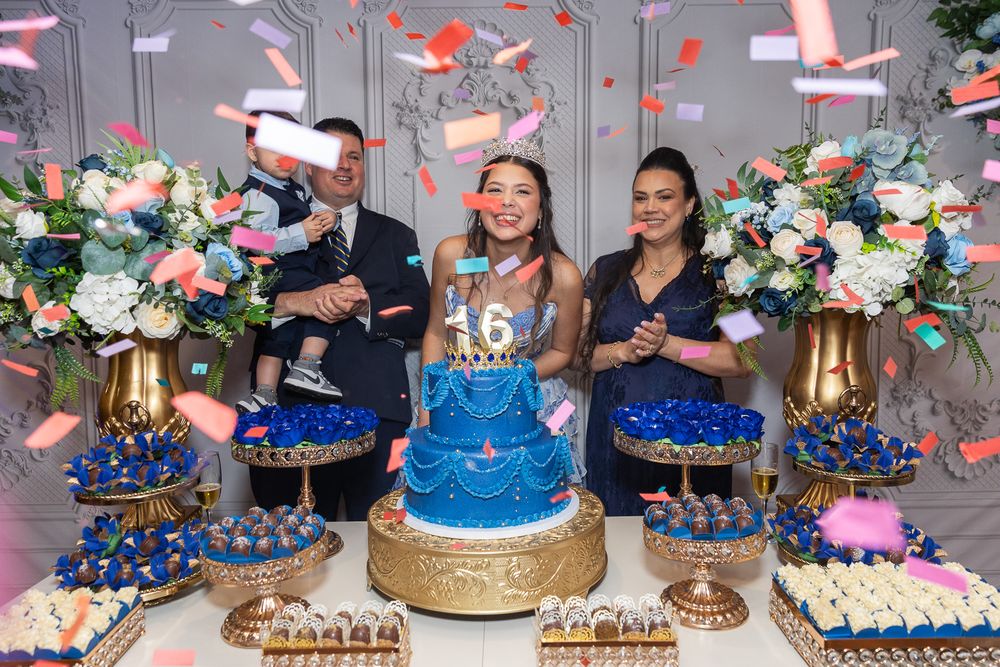 A sweet 16 birthday celebration with a blue tiered cake, dessert table, confetti, and three people posing joyfully.