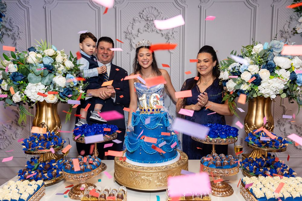 Teen girl celebrates quinceañera with family beside a blue tiered cake surrounded by colorful confetti and floral arrangements.