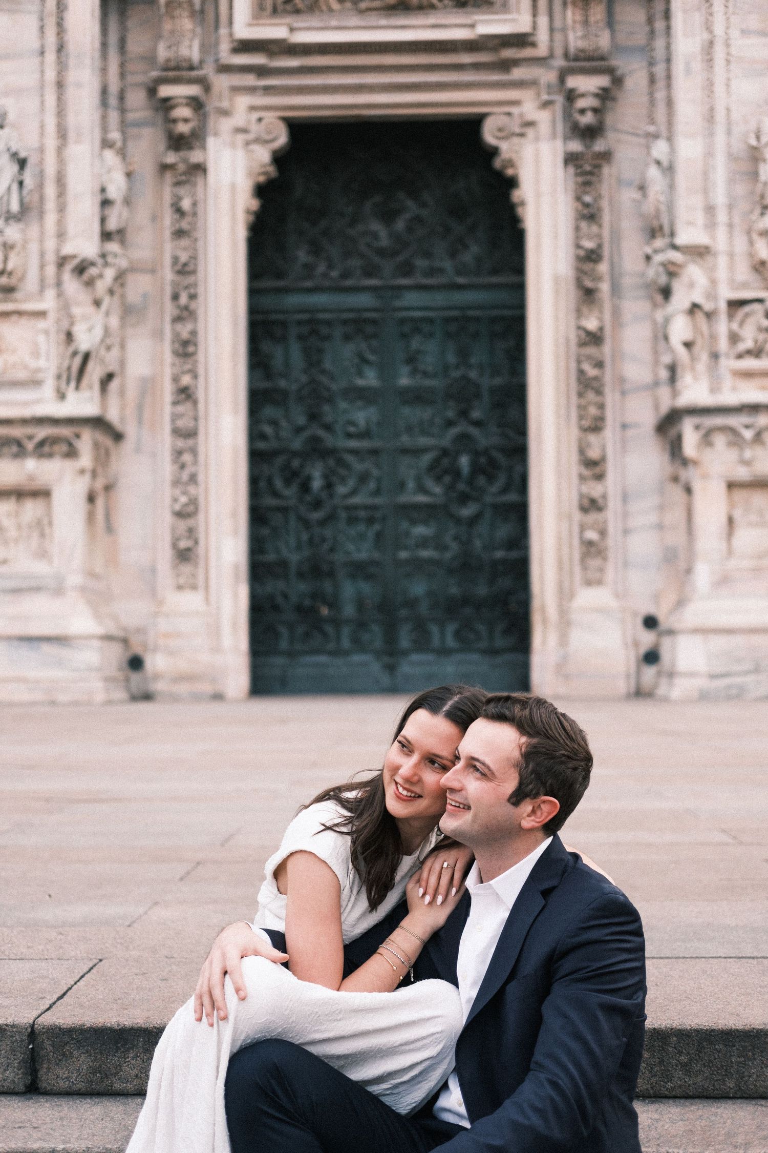 Couple sitting on steps in front of Milan Cathedral, smiling and enjoying a romantic moment together.