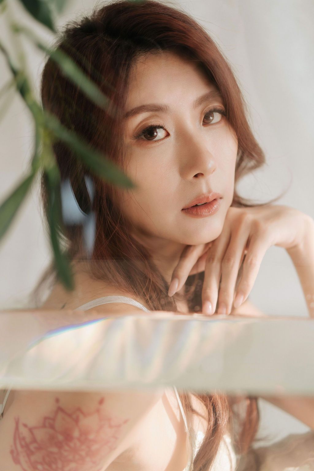 Young Asian woman with brown hair and tattoo poses gracefully against a soft white background with green plants.