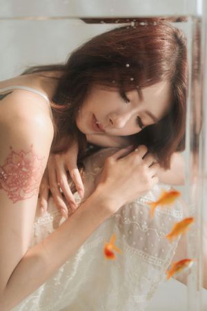 Young Asian woman in white lace dress with floral tattoo on shoulder, leaning near fish tank with goldfish.