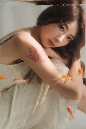 Young woman in white lace dress with floral tattoo on arm, surrounded by goldfish in dreamy underwater-style portrait.