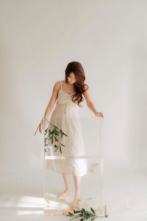 Woman in white slip dress holding a clear acrylic box with green branches and orange petals on white background.