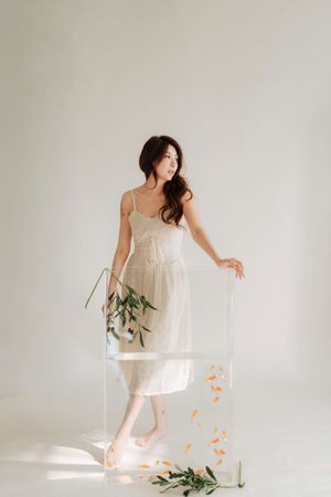 Woman in white midi dress standing beside a clear acrylic box with orange petals and green branches on white background.
