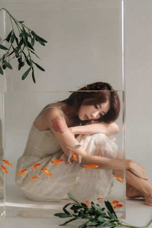 Young woman in beige dress sits inside glass tank with goldfish and olive branches in artistic photo.