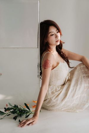 Young woman with floral tattoo in beige slip dress sits against white wall with olive branch.