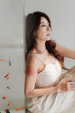 Young Asian woman in white dress with floral tattoo sits beside goldfish tank in bright minimalist setting.