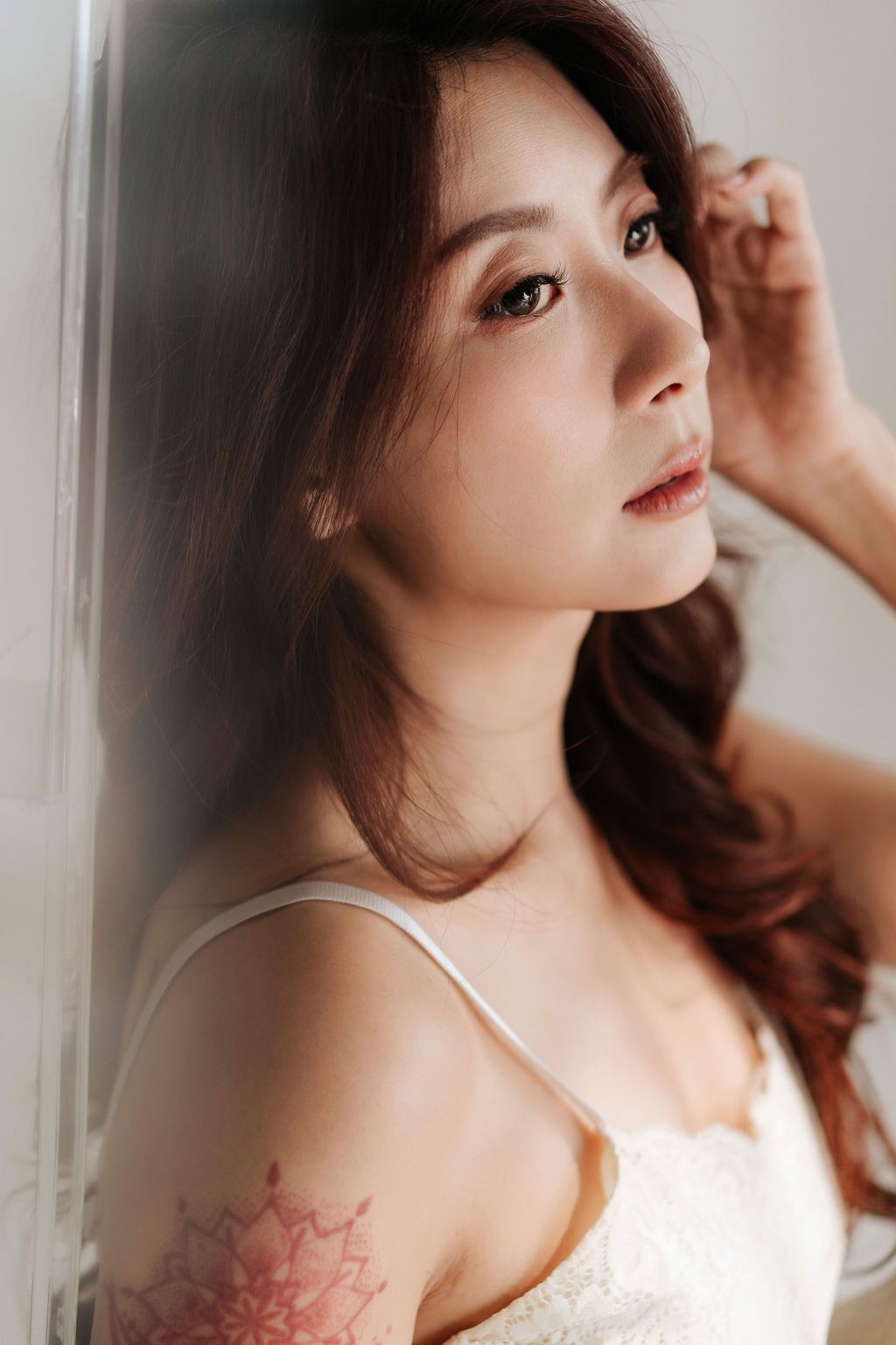 Young Asian woman with long brown hair and floral tattoo on shoulder poses thoughtfully against a white wall.