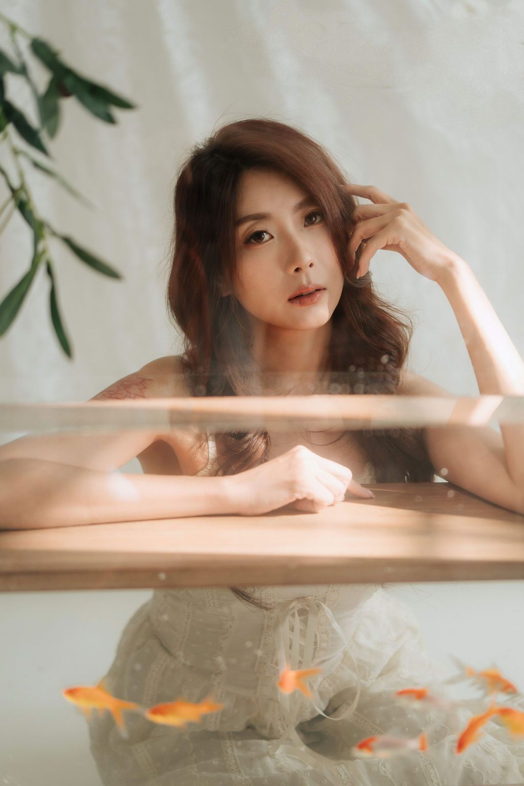 A young Asian woman in a white dress leans on a glass fish tank with goldfish, posing elegantly in a soft-lit studio.