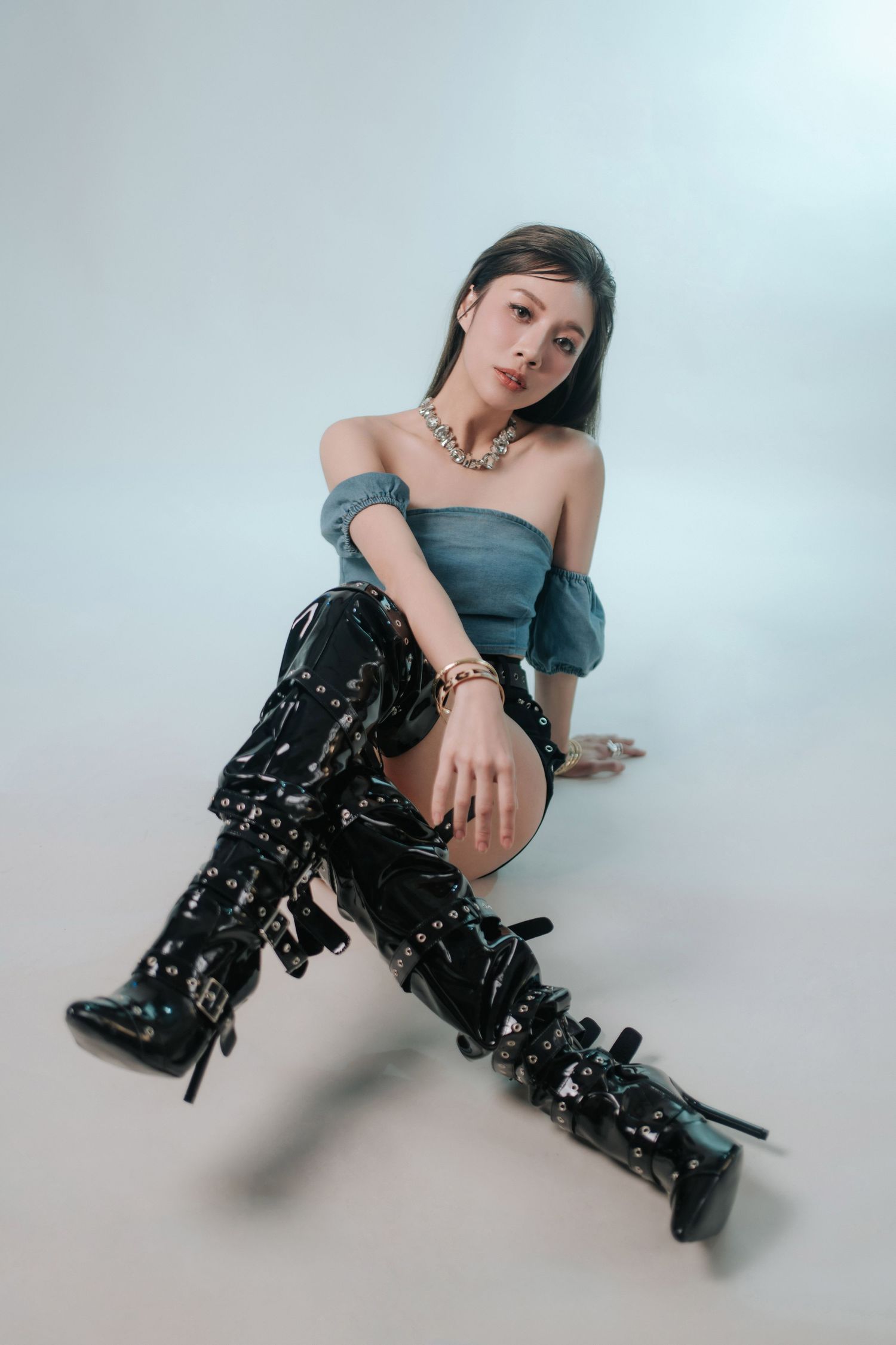 Young woman in denim off-shoulder top, black patent thigh-high boots, and gold jewelry poses on white studio floor.