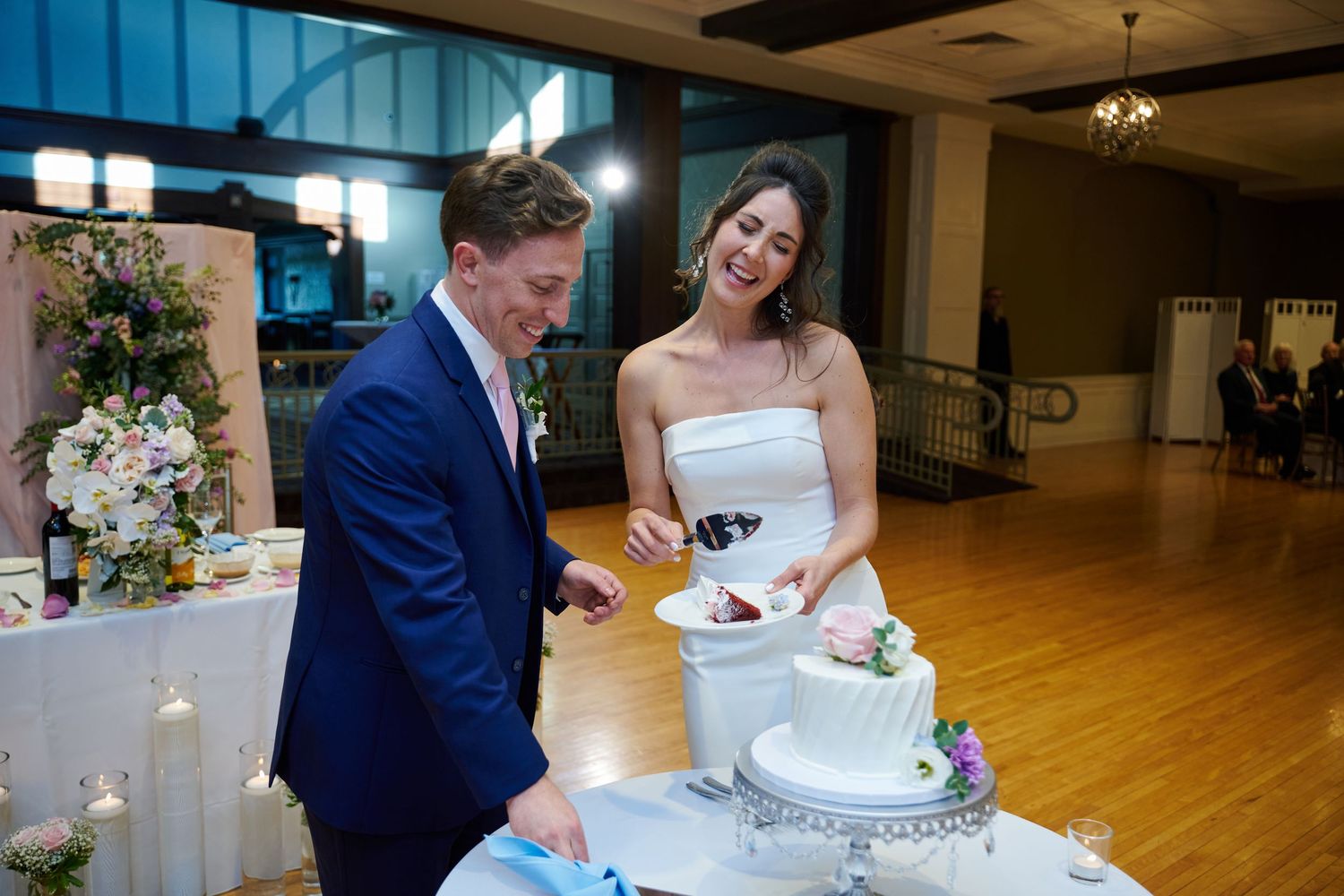 Bride and groom smiling while cutting their white wedding cake at an elegant indoor reception venue.