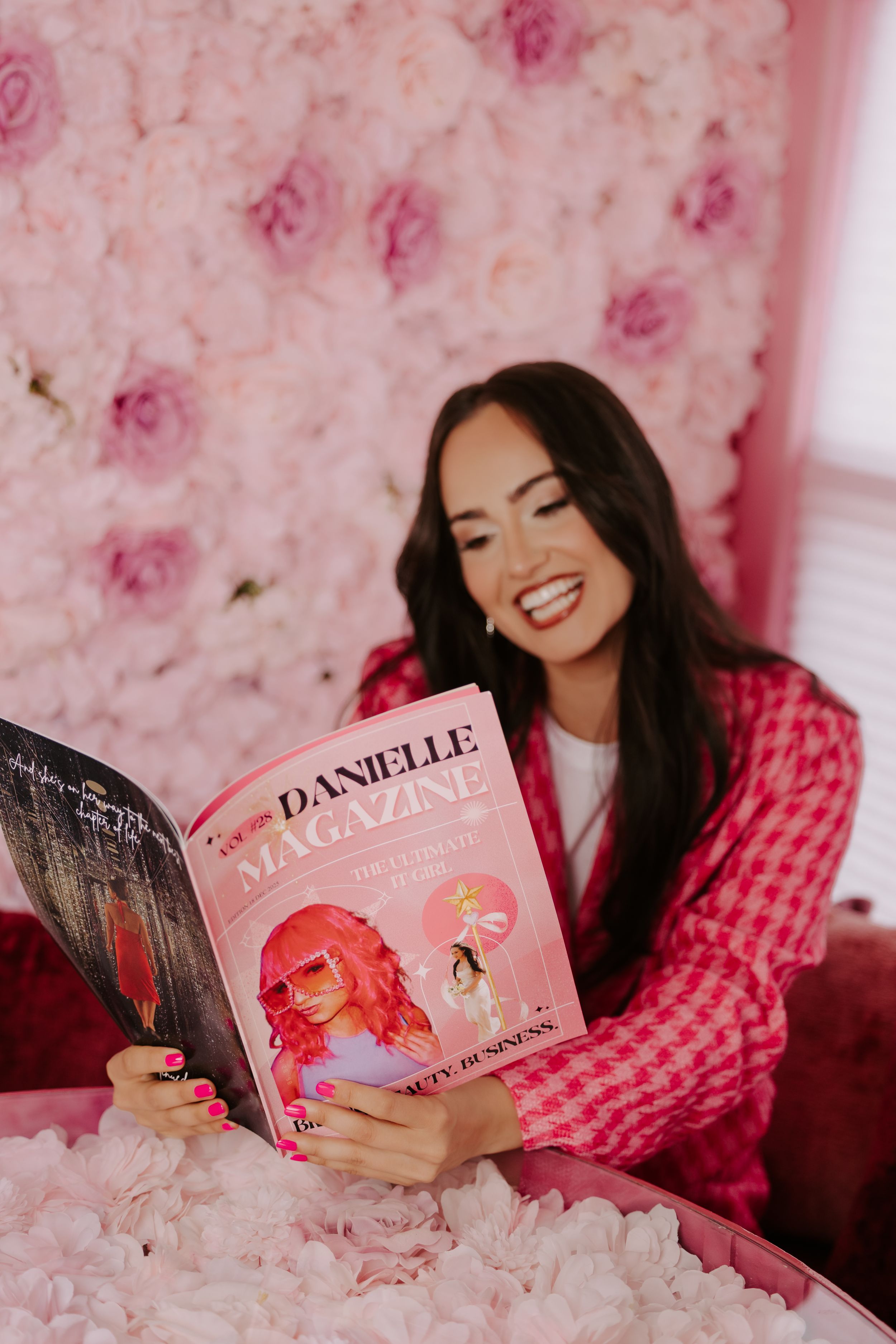 Woman in pink blazer reading Danielle Magazine against a floral pink backdrop, smiling joyfully.