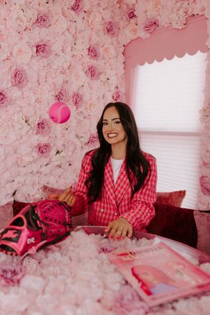 Woman in pink houndstooth blazer poses at floral pink table with magazine and handbag against rose wall.