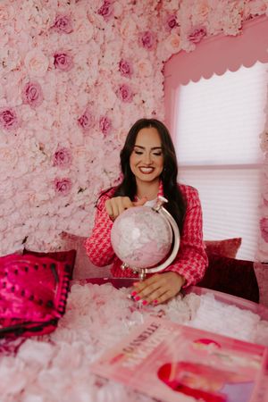 Woman in pink houndstooth blazer smiling while holding a pink globe at a floral rose wall backdrop.