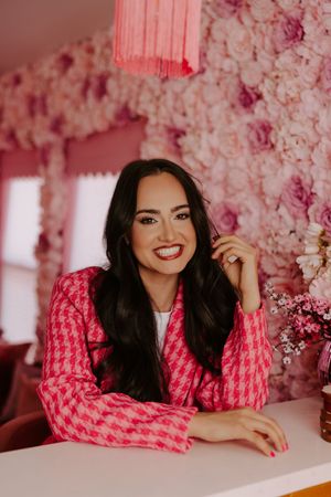 Smiling woman in pink houndstooth blazer sits at white table against floral pink wall backdrop.