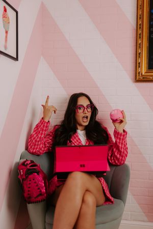 Woman in pink tweed suit sits in chair with pink laptop, holding macaron and pointing finger up.