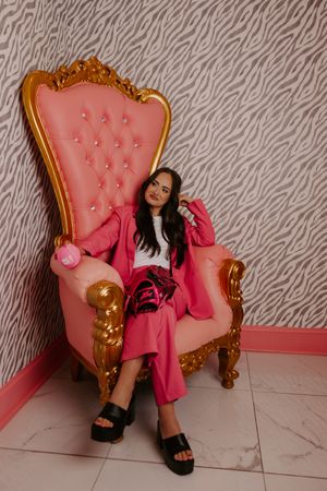 Woman in pink suit reclining on a gold and pink tufted throne chair against zebra wallpaper.