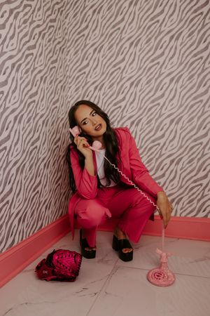 Woman in hot pink pantsuit crouching on floor holding a vintage pink rotary phone by zebra wallpaper.