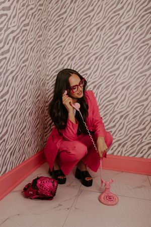 Woman in pink suit crouching with vintage pink rotary phone against zebra print wallpaper.