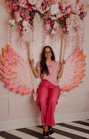Woman in pink outfit sitting on floral swing with angel wings decoration and flowers.
