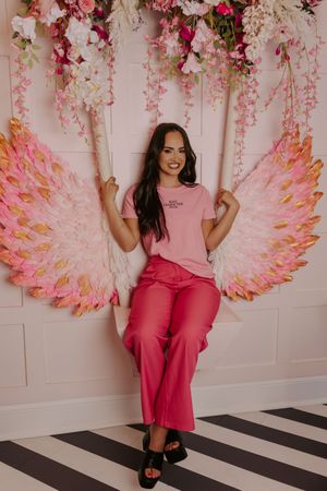 Woman in pink outfit poses on decorative swing with large angel wings and floral arrangement.