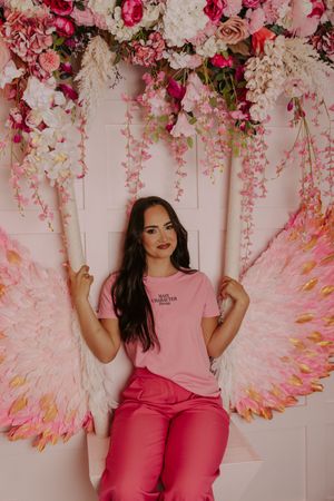 Woman in pink t-shirt and pants posing on swing with large angel wings and floral backdrop.
