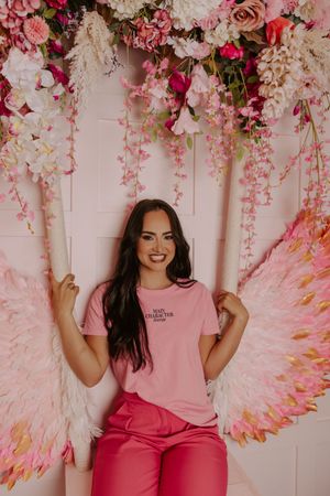 Smiling woman in pink t-shirt and pants sitting on a floral swing with angel wings backdrop.