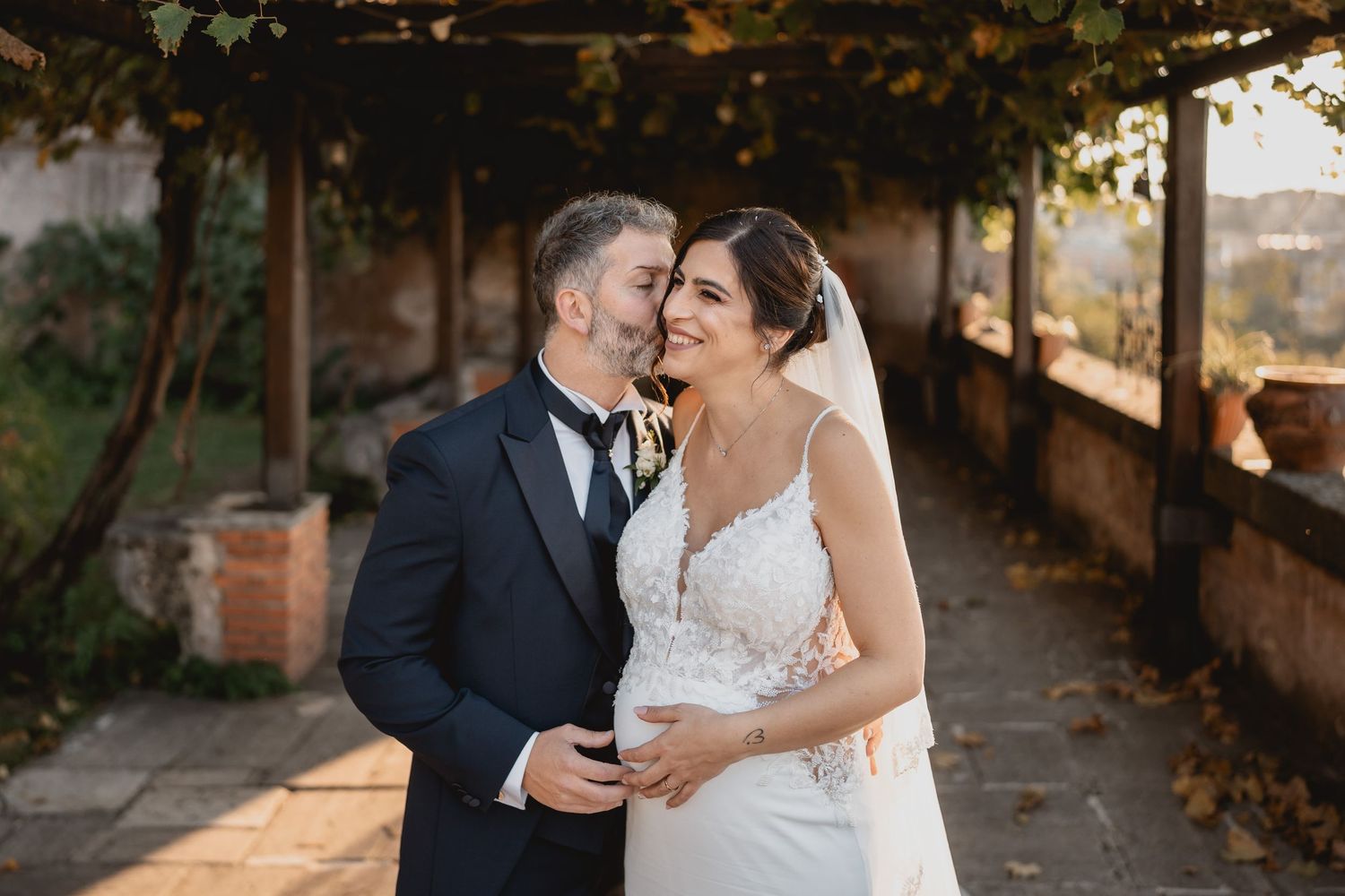 A couple in wedding attire share a romantic moment under a vine-covered pergola at a rustic outdoor venue.