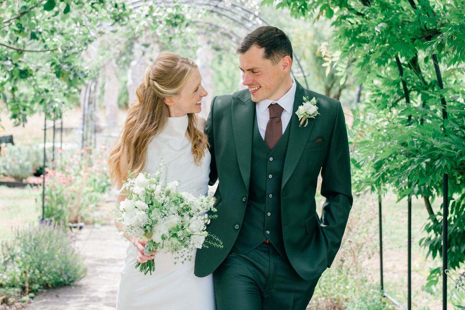 A bride and groom smile at each other in a lush garden, she holds white flowers, he wears a green suit in the Tangled Garden