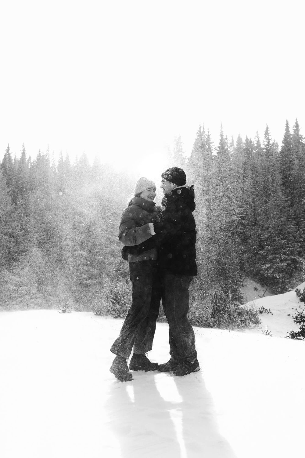 Black and white photo of a couple embracing in a snowy winter landscape with snow-covered pine trees.