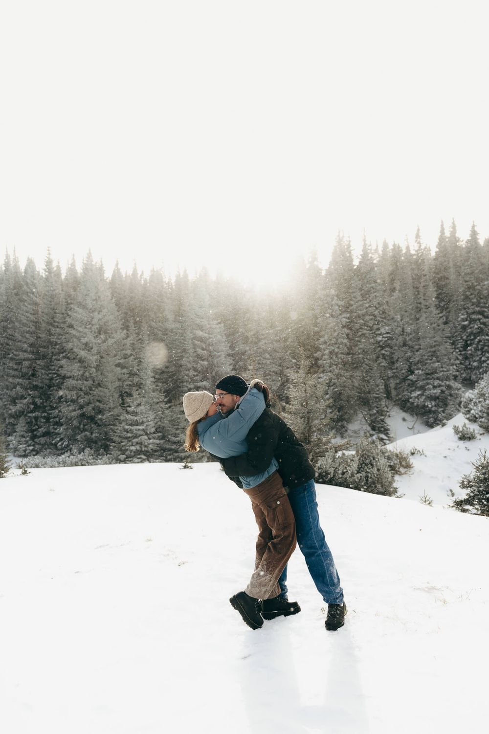 Couple embracing and kissing in a snowy winter landscape surrounded by snow-covered evergreen trees.