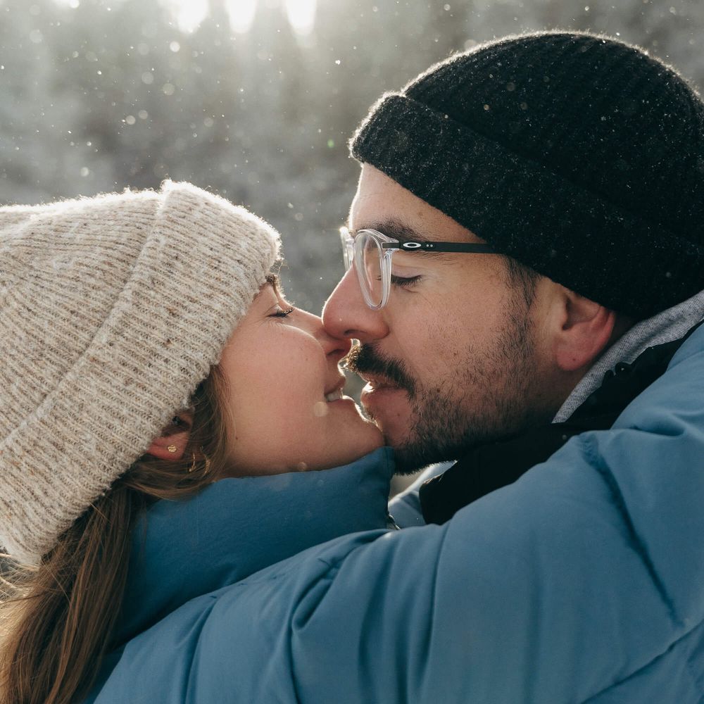 Couple sharing a romantic kiss in a snowy winter setting, wearing cozy hats and winter jackets.