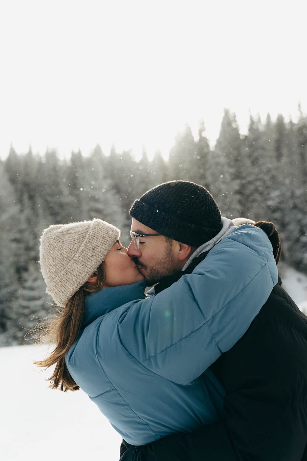 Couple sharing a romantic kiss in snowy winter landscape, wearing warm hats and jackets with snow-covered pine trees.